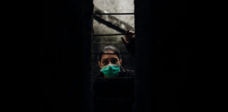 What COVID-19 Has to Teach Us About Psychiatric Oppression woman in medical mask peers out of darkness