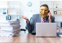 BMJ: 20% of Health Research Is Fraudulent Photo of a man in a suit wearing a mask at a desk with a computer and a tall pile of paperwork