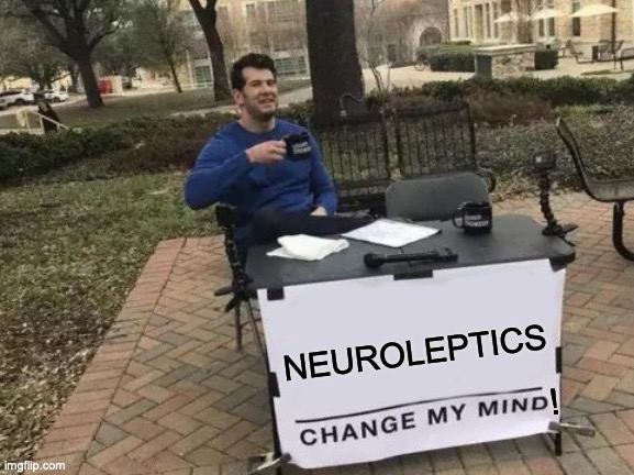 An image of a man behind a table with a cup of coffee. The table has a sign that says "Neuroleptics Change my mind"