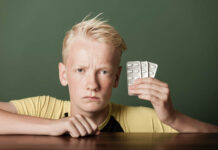 Risk of Depression Spikes When Kids Take Ritalin Scowling teenage boy holding up pills sealed in blister packs in his hand as he leans on a wooden table with an intense stare