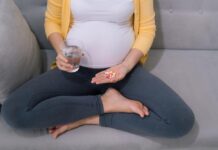 Dubious Science: Downplaying the Risks of Antidepressants in Pregnancy Photo of pregnant person holding a glass of water and a handful of pills. Sitting cross-legged on couch. We cannot see her face.
