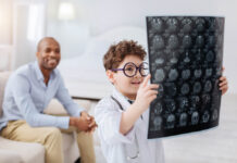 Critical Psychiatry Textbook, Chapter 3: Are Psychiatric Disorders Detectable in a Brain Scan? Photo of a young White boy with glasses looking at a giant sheet of brain imaging results, with a Black man smiling happily behind him