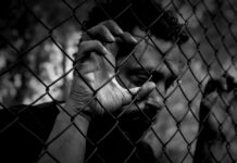 The Hidden Injuries of Oppression Black and white photo of a man looking sad with his fingers clinging to a chain-link fence