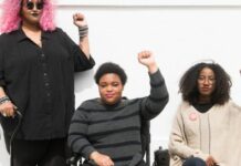 Therapist Trainings Needed on Disability to Counter Neoliberalism in Mental Healthcare orso level photo of three Black and disabled folx (a non-binary person holding a cane, a woman in a power wheelchair, and a woman on a folding chair) raising their fists on the sidewalk in front of a white wall. (Credit: Disabled and Here.)
