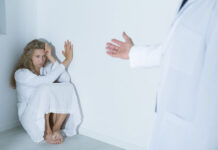 An Unacknowledged Role of Doctors: Suppressing Women A young woman crouches in distress in a corner, while a doctor's hand reaches out to her. Everything is white