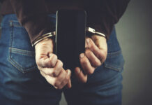 Psychiatric Detentions Rise 120% in First Year of 988 Photo depicting a close-up of hands handcuffed behind someone's back, holding a cell phone