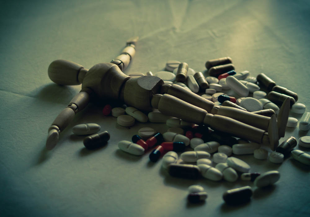 An artist's posable mannequin lies flat amongst a pile of pills, dark lighting