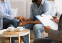De-privatizing Our Relationships A black couple holds hands out of focus in background. In foreground, a therapist's hands on a clipboard taking notes.