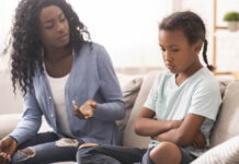 The Psychiatric Peddlers in Your Schools A Black mother and daughter sit on the couch. Mother is explaining something to a sullen-looking girl.
