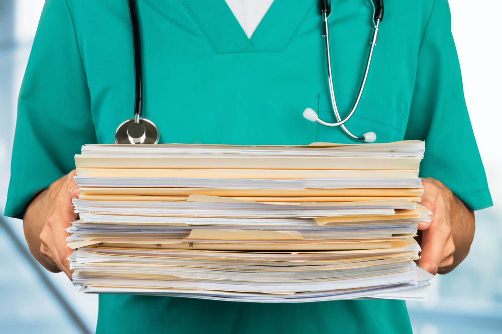 Closeup shot of a figure in blue/green scrubs holding a pile of paperwork and folders