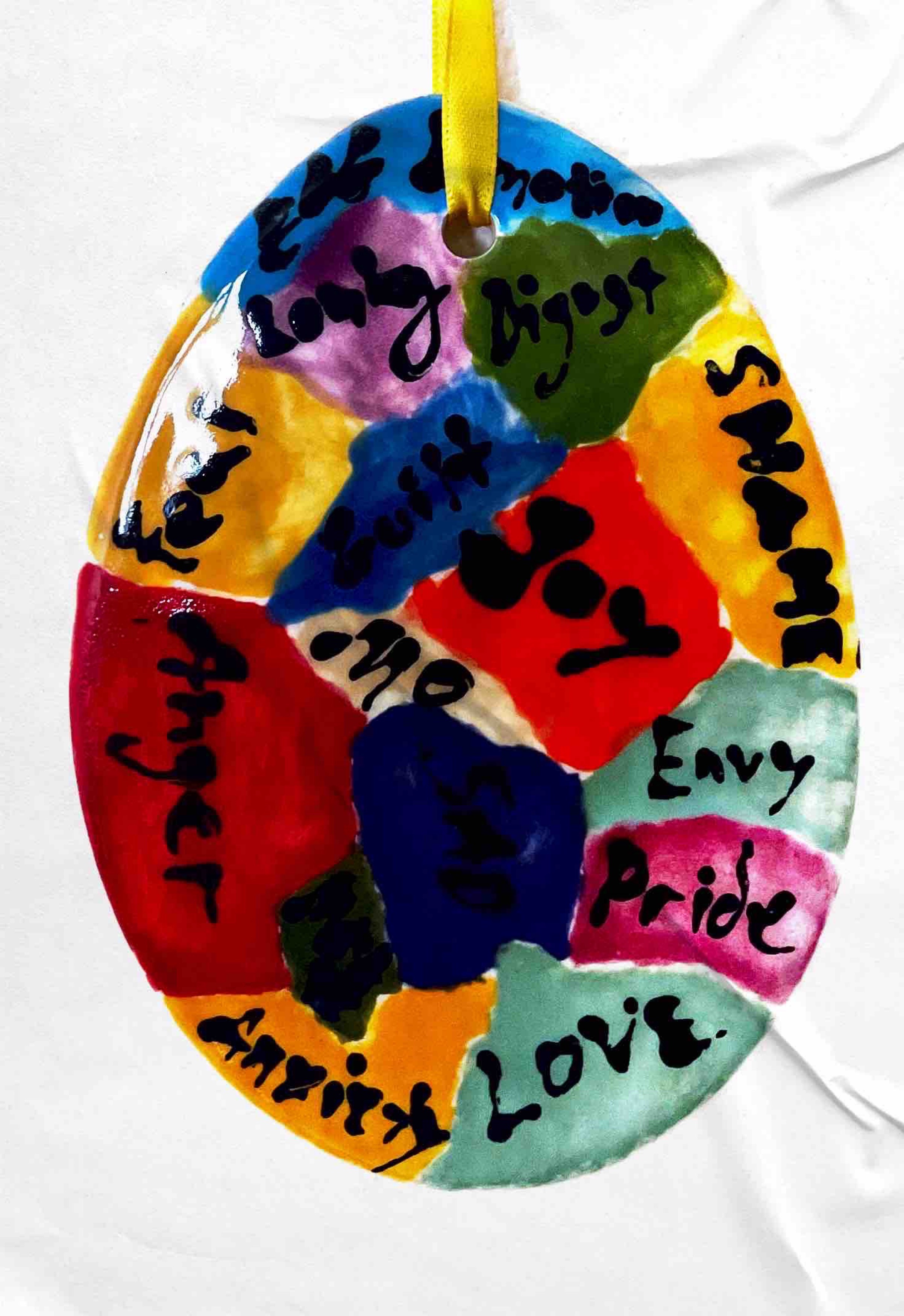 egg-shaped piece glazed with fields of color with words in black such as "Joy," "Envy,", "Anger," "Pride" and "Love," suspended on a yellow cord, white background