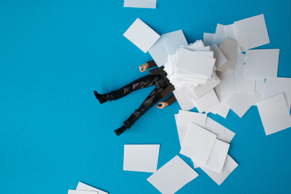 Miniature photography. A man's legs are visible under a pile of paperwork on a blue background.