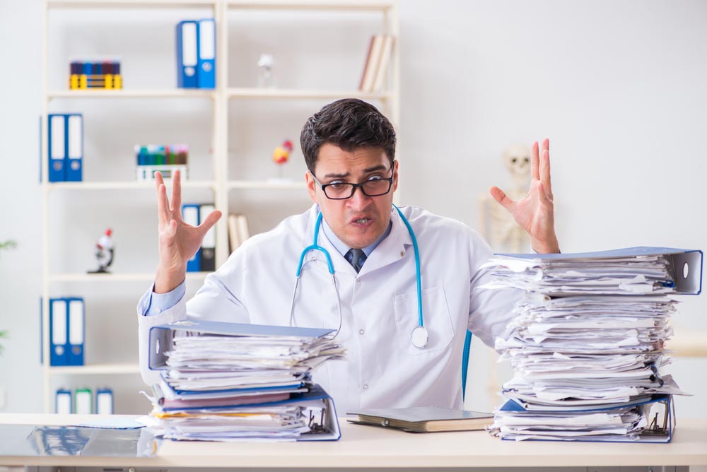 Doctor looking frustrated raising hands behind a desk with piles of paper