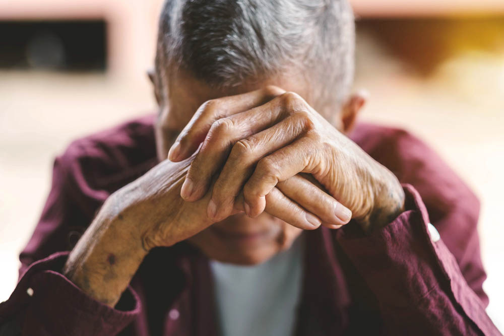 An older man holds his hands together obscuring his face, head dropped in sadness