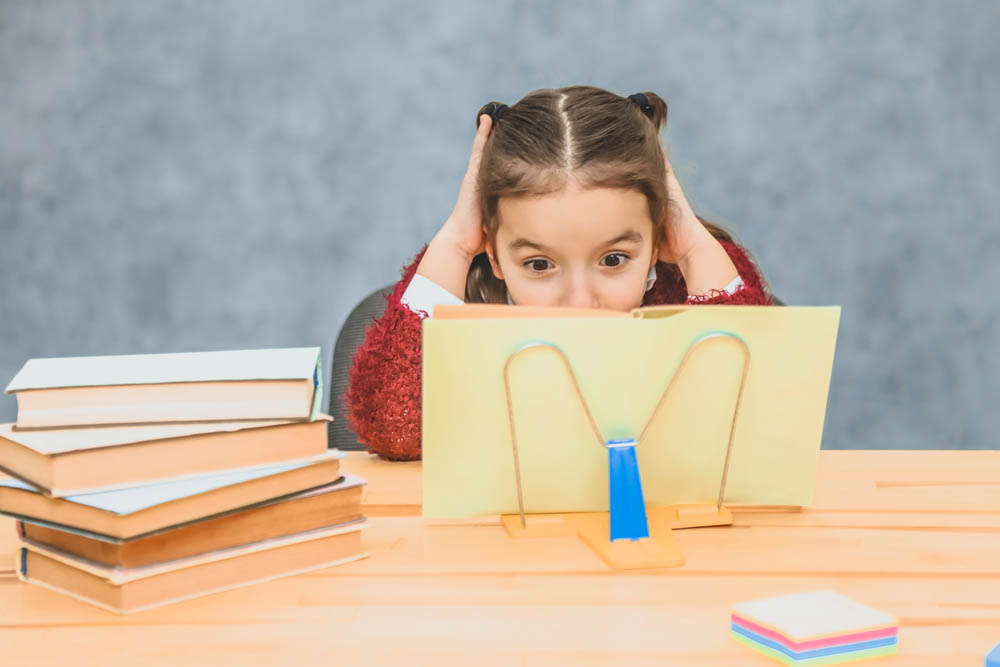 A sweet girl schoolgirl on a gray background. Works homework by reading a book in a yellow cover.