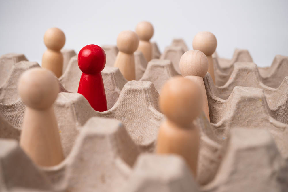 Macro photography of wooden blocks representing people. One red one stands among wood-colored ones.