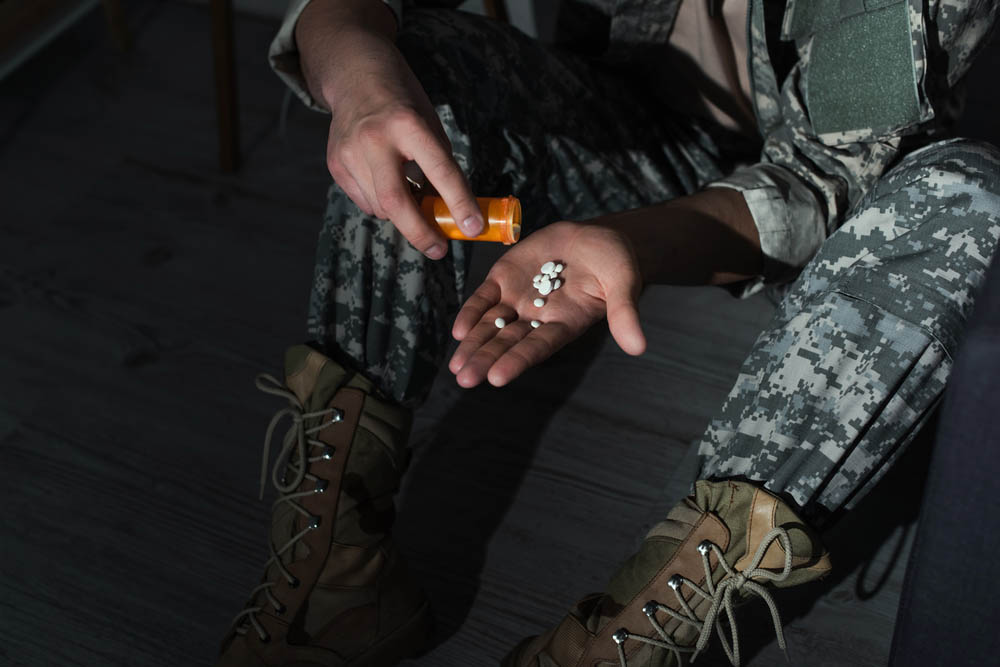 A hand holding pills from a pill bottle. Legs in military camo are visible