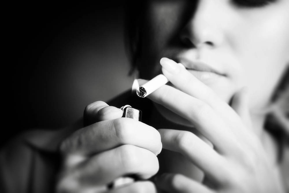 B&W photo of a young woman smoking. Close-up on the cigarette being lit by a lighter; her face is out of focus