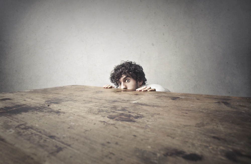 A man hiding behind a large wooden table, only his head and hands showing
