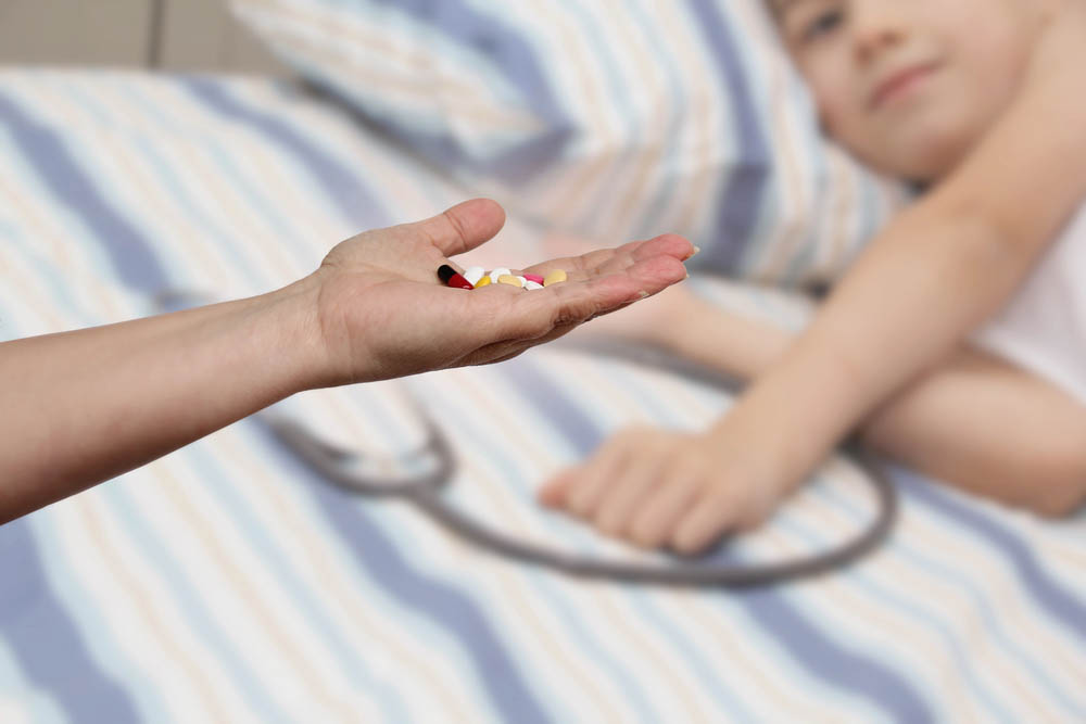 A hand holds out pills. A child is out of focus in the background.