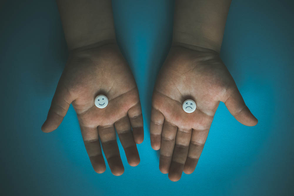 Two hands holding pills, one with a smiley face, the other with x's for eyes and a frown