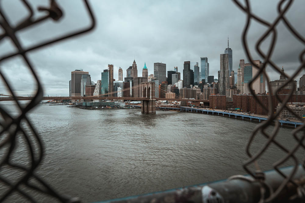 View of New York City skyline through a chain-link fence, featuring the Brooklyn Bridge and skyscrapers under a cloudy sky.
